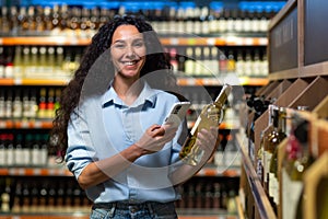 Portrait of happy and smiling woman shopper in supermarket, latin american woman choosing alcohol wine using app on