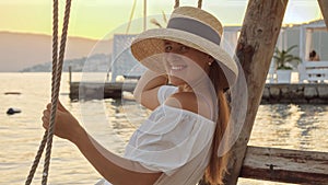 Portrait of happy smiling woman in straw hat sitting on the swing against sunset over the sea beach