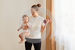 Portrait of happy smiling woman wearing white t shirt holding baby daughter in hands and training biceps and triceps with raising