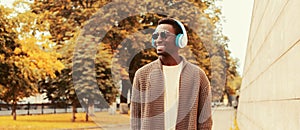 Portrait of happy smiling young african man in headphones listening to music wearing brown knitted cardigan in the city park