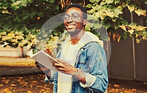 Portrait of happy smiling young african man student reading a book wearing eyeglasses in autumn city park