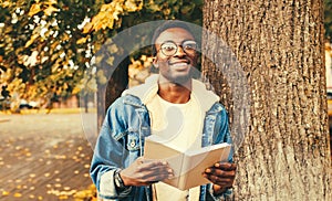 Portrait of happy smiling young african man student reading a book wearing eyeglasses in autumn city park