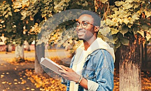 Portrait of happy smiling young african man student reading a book wearing eyeglasses in autumn city park