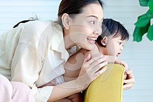 Portrait of happy smiling young Asian mother hugging her toddle baby girl daughter on yellow sofa in living room at home, parent