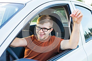 Portrait of happy smiling young man, buyer sitting in his new car and showing keys outside dealer office. Personal