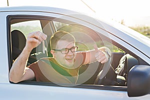 Portrait of happy smiling young man, buyer sitting in his new car and showing keys outside dealer office. Personal