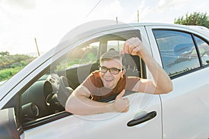 Portrait of happy smiling young man, buyer sitting in his new car and showing keys outside dealer office. Personal