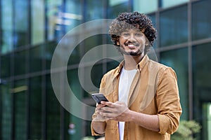 Portrait of a happy and smiling young Muslim man standing on a city street using a mobile phone and looking confidently