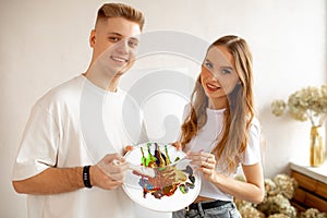 Portrait of happy smiling young pretty couple stand closely, posing, holding watercolor palette and brushes. Art therapy