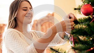 Portrait of happy smiling young woman hanging beautiful bauble on branch of Christmas tree at morning