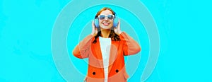 Portrait of happy smiling young woman in headphones listening to music wearing red jacket on blue background