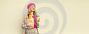 Portrait of happy smiling young woman holding grocery shopping paper bag with long white bread baguette and cup of coffee on gray