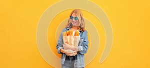 Portrait of happy smiling young woman holding grocery shopping paper bag with long white bread baguette on orange background