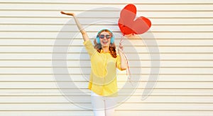 Portrait of happy smiling young woman listening to music in headphones holding bunch of red heart shaped balloons on white