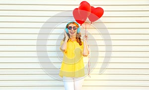 Portrait of happy smiling young woman listening to music in headphones holding bunch of red heart shaped balloons on white