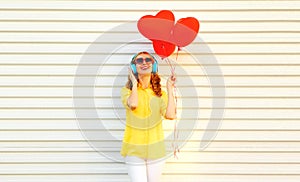 Portrait of happy smiling young woman listening to music in headphones holding bunch of red heart shaped balloons on white