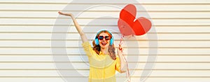 Portrait of happy smiling young woman listening to music in headphones holding bunch of red heart shaped balloons on white