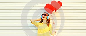 Portrait of happy smiling young woman listening to music in headphones holding bunch of red heart shaped balloons on white