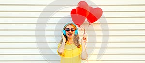 Portrait of happy smiling young woman listening to music in headphones holding bunch of red heart shaped balloons on white