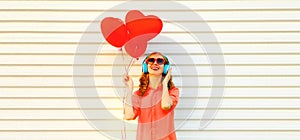 Portrait of happy smiling young woman listening to music in headphones holding bunch of red heart shaped balloons on white