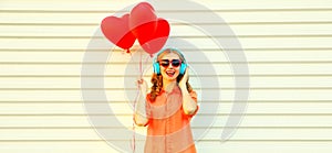 Portrait of happy smiling young woman listening to music in headphones holding bunch of red heart shaped balloons on white
