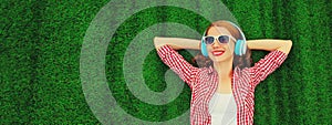 Portrait of happy smiling young woman listening to music in headphones relaxing lying on the grass background in summer park