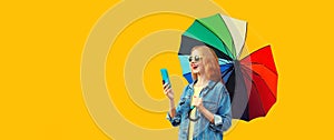 Portrait of happy smiling young woman looking at smartphone with colorful umbrella isolated on yellow background, blank copy space