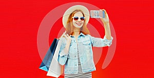 Portrait of happy smiling young woman with shopping bags taking selfie by smartphone wearing summer straw hat and denim jacket on