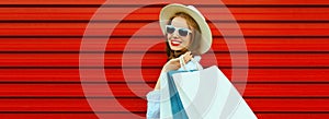 Portrait of happy smiling young woman with shopping bags wearing straw hat and shirt on colorful red background