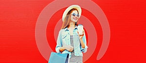 Portrait of happy smiling young woman with shopping bags wearing summer straw hat, denim jacket on red background