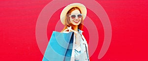 Portrait of happy smiling young woman with shopping bags wearing summer straw hat, denim jacket on red background