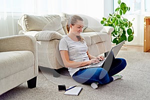 Portrait of happy smiling young woman sitting on floor and typing on laptop at home, copy space