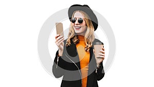 Portrait of happy smiling young woman with smartphone and cup of coffee wearing black coat, round hat, sunglasses isolated on