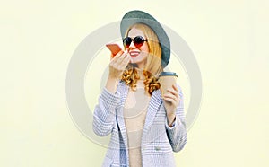 Portrait happy smiling young woman with smartphone using voice command recorder or calling, wearing coat jacket, round hat on wall