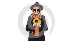 Portrait of happy smiling young woman with smartphone wearing black coat, round hat, sunglasses isolated on white background