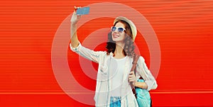 Portrait of happy smiling young woman taking selfie by phone wearing summer straw hat, backpack on red background