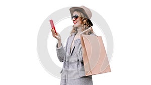 Portrait of happy smiling young woman taking selfie with smartphone holding shopping bags wearing coat, round hat isolated on