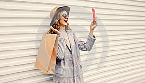 Portrait of happy smiling young woman taking selfie with smartphone holding shopping bags wearing coat, round hat on white