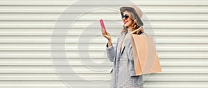 Portrait of happy smiling young woman taking selfie with smartphone holding shopping bags wearing coat, round hat on white