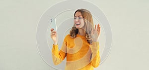 Portrait of happy smiling young woman taking selfie with smartphone or talking on video call waving hand wearing casual knitted