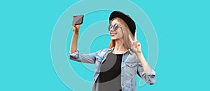 Portrait of happy smiling young woman taking selfie with smartphone wearing black round hat isolated on blue background