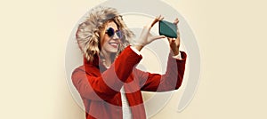 Portrait of happy smiling young woman taking selfie with smartphone wearing red jacket with fur hood