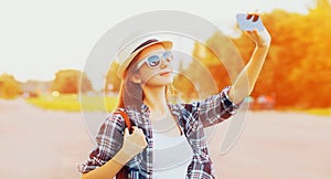 Portrait of happy smiling young woman taking selfie by smartphone wearing summer straw hat, backpack in summer park