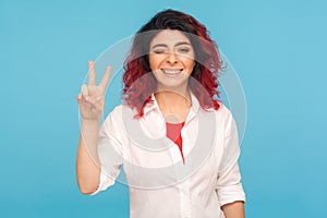 Portrait of happy successful hipster woman with fancy red hair winking playfully to camera and showing victory gesture