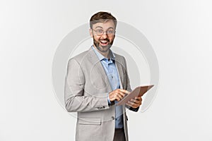 Portrait of happy and surprised bearded businessman in grey suit and glasses, holding digital tablet, smiling amazed at