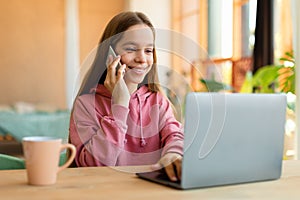Portrait of happy teen girl talking on cellphone and typing on laptop keyboard, sitting at desk, talking with friends