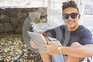 Portrait of happy teenage boy in sunglasses, using digital tablet while sitting on boundary wall beside fence. Smiling confident