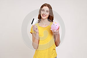 Portrait of happy teenager girl with brown hair wearing yellow casual style T- shirt holding credit card and piggy bank, saving