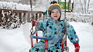 Portrait of happy toddler boy enjoying a winter swing ride in the park, surrounded by snow-covered trees and beautiful winter