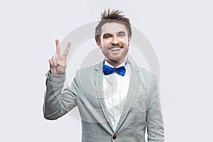 Portrait of happy toothy smiley handsome bearded man in casual grey suit, blue bow tie standing and looking at camera with victory